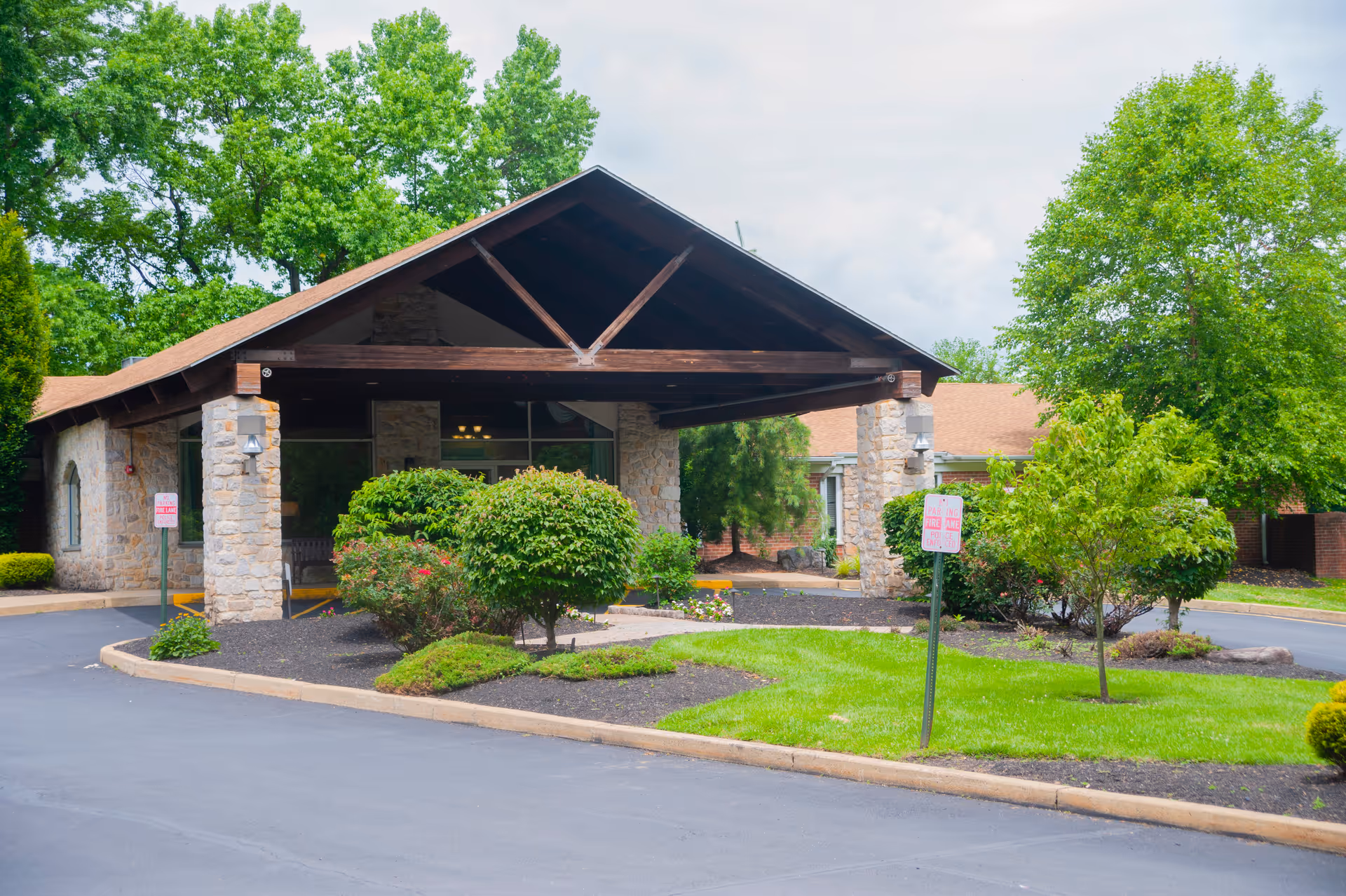 Front entrance of a stone-faced senior living building with a wooden covered porte-cochere, manicured shrubs, and a circular driveway.
