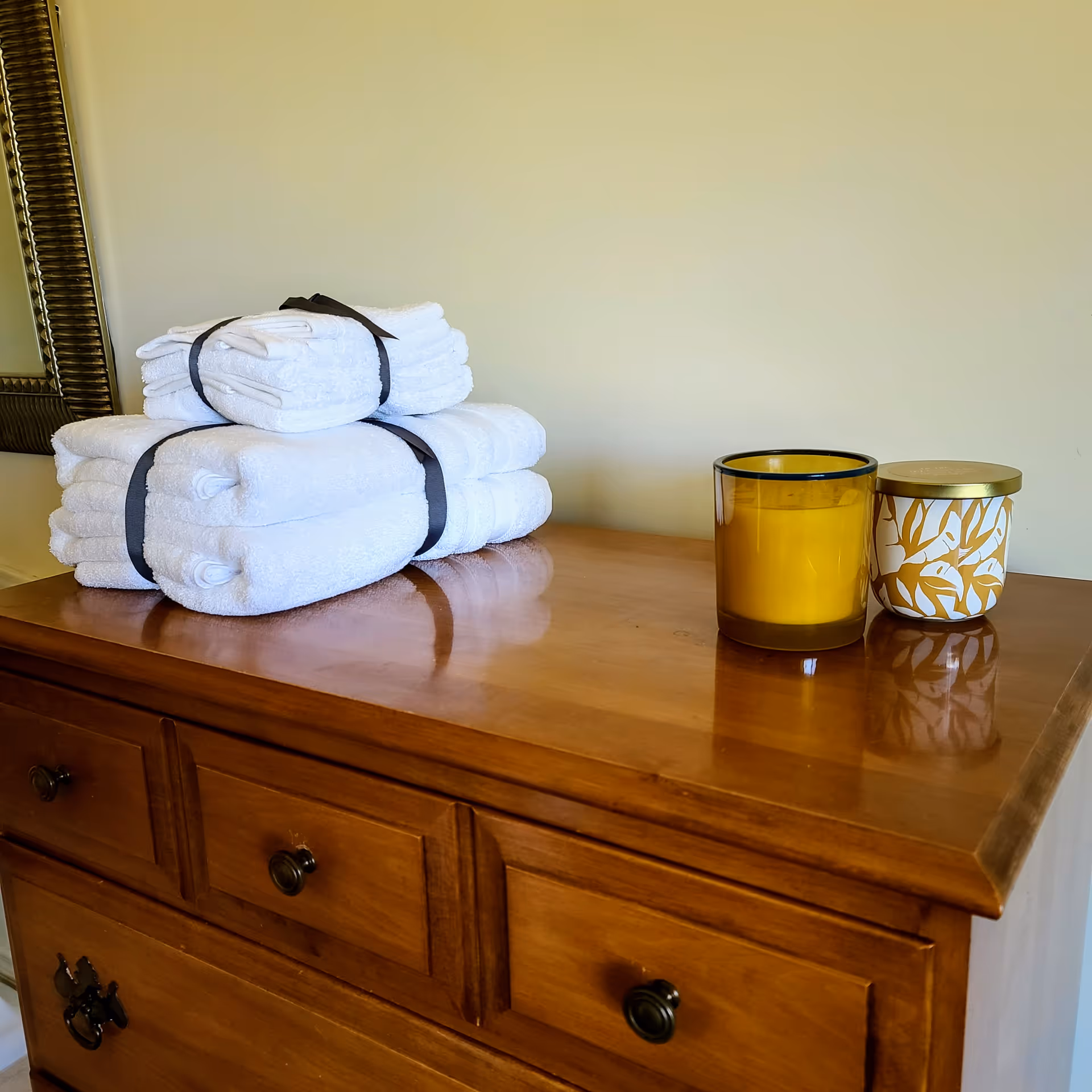 A wooden dresser with three drawers, on top of which are neatly stacked white towels tied with black ribbons, alongside two decorative candles, one in a yellow glass holder and the other in a white and gold patterned container.