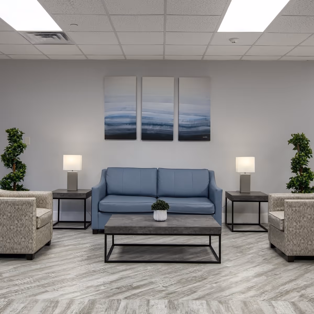 A modern nursing center lounge area with a blue sofa centered between two beige armchairs. There are two side tables with lamps on either side of the sofa, a rectangular coffee table with a small potted plant in front, and two tall green plants flanking the seating arrangement. Above the sofa, three abstract blue and gray paintings are hung on a light gray wall. The floor has a light wood pattern, and the ceiling has white tiles with fluorescent lighting.