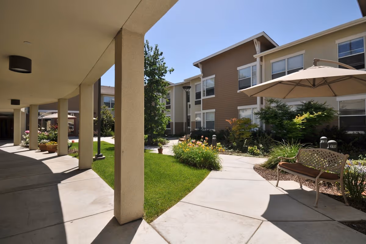 Outdoor courtyard area of a senior living facility with a covered walkway supported by columns, green grass, flower beds, a bench with a cushion under a large patio umbrella, and a multi-story building in the background under a clear blue sky.