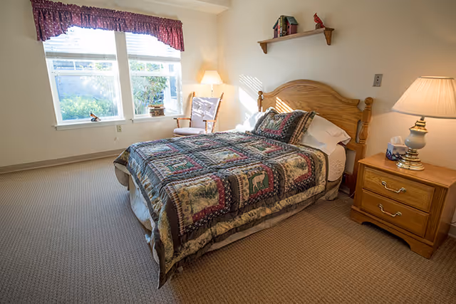 A cozy bedroom with a wooden bed frame and a patchwork quilt featuring animal designs. There is a wooden nightstand with a lamp and a box of tissues on it. A wooden rocking chair is placed near a window with white blinds and a red valance. A small shelf with decorative items is mounted on the wall above the bed.