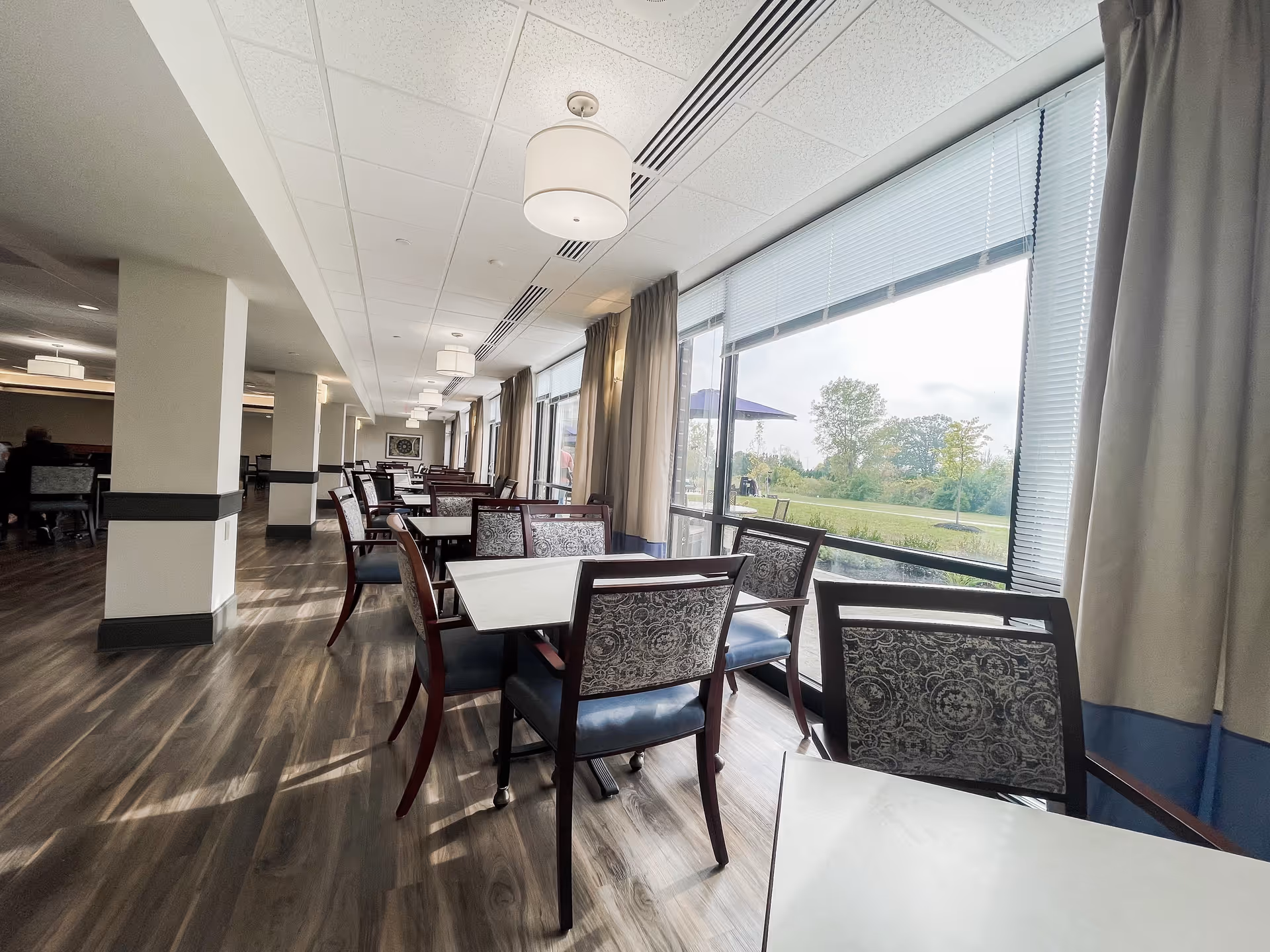 A spacious dining room with multiple tables and chairs arranged in rows. Large windows on one side allow natural light to fill the room, showing a view of greenery outside. The floor has a wood-like finish, and ceiling lights hang evenly spaced along the length of the room.