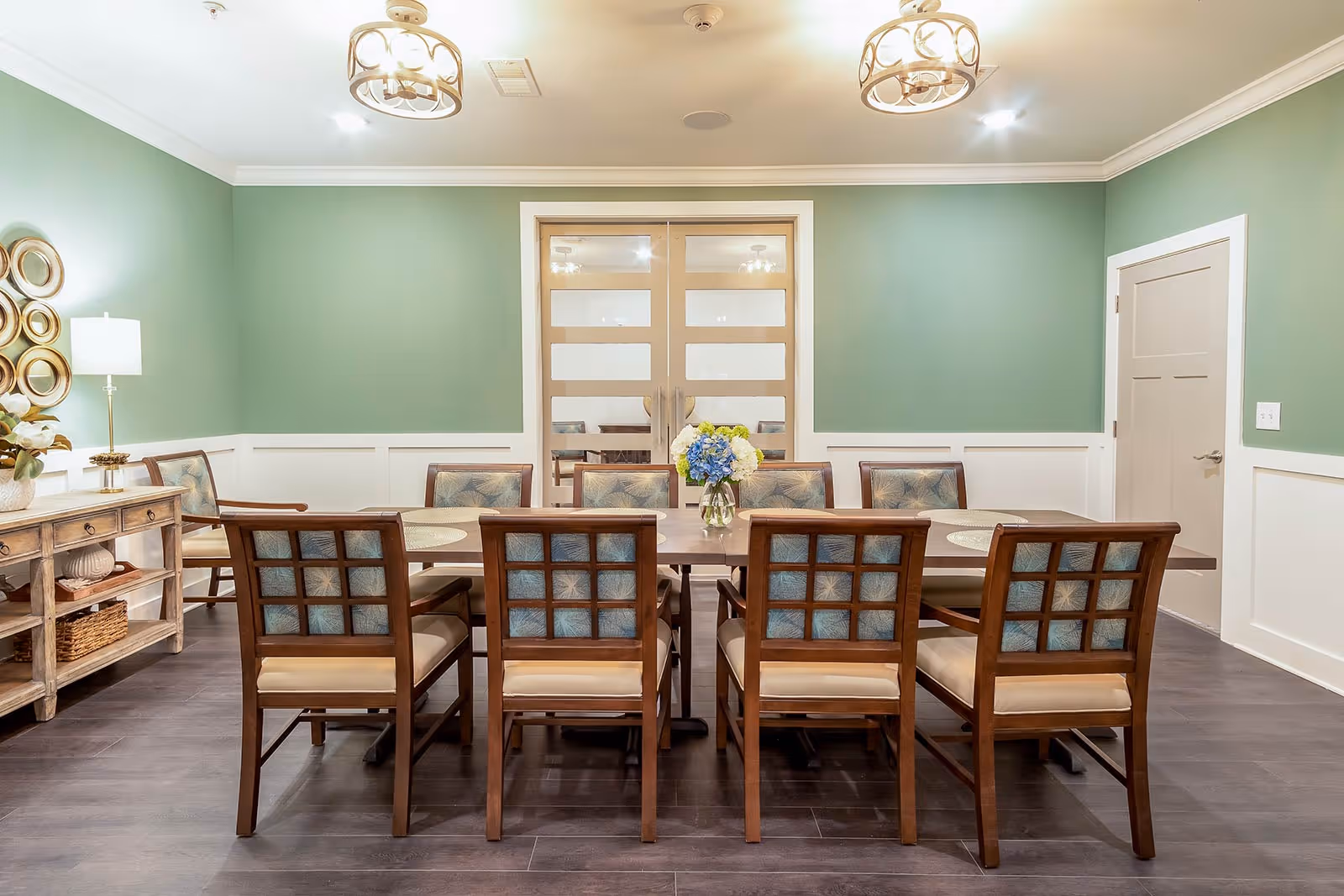 A dining room with a long wooden table surrounded by eight cushioned chairs, green walls, and decorative ceiling lights.