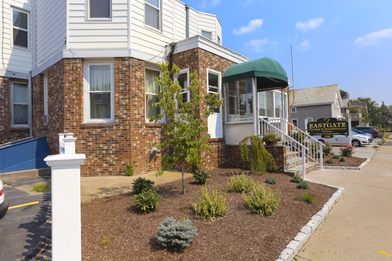 Exterior view of Eastgate Nursing & Rehabilitation Center showing a brick and white siding building with a green awning over the entrance. There is a small landscaped area with various plants and shrubs in front of the building, a sidewalk, and a parking area with several cars. A sign near the entrance displays the facility's name.