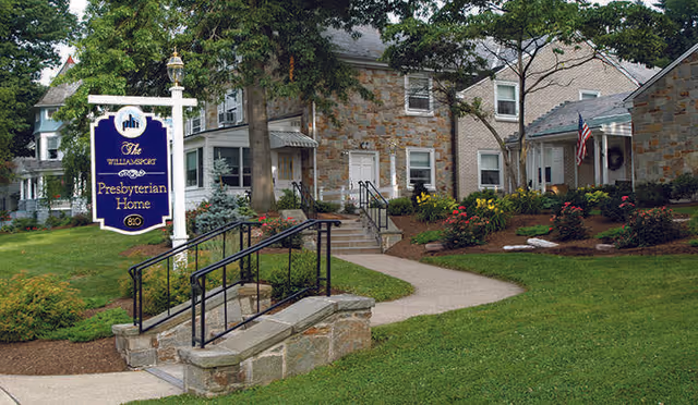 Exterior view of the Presbyterian Home at Williamsport, showing a stone and brick building with a landscaped garden, a pathway, and a signpost with the facility's name. There are trees, flowers, and an American flag near the entrance.