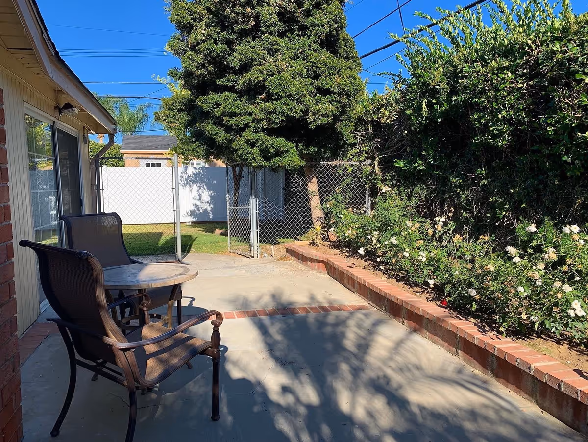 Outdoor patio area with two brown mesh chairs and a round table on a concrete surface next to a building. There is a brick border garden bed with green bushes and white flowers on the right side. A large tree and a chain-link fence with a gate are visible in the background under a clear blue sky.