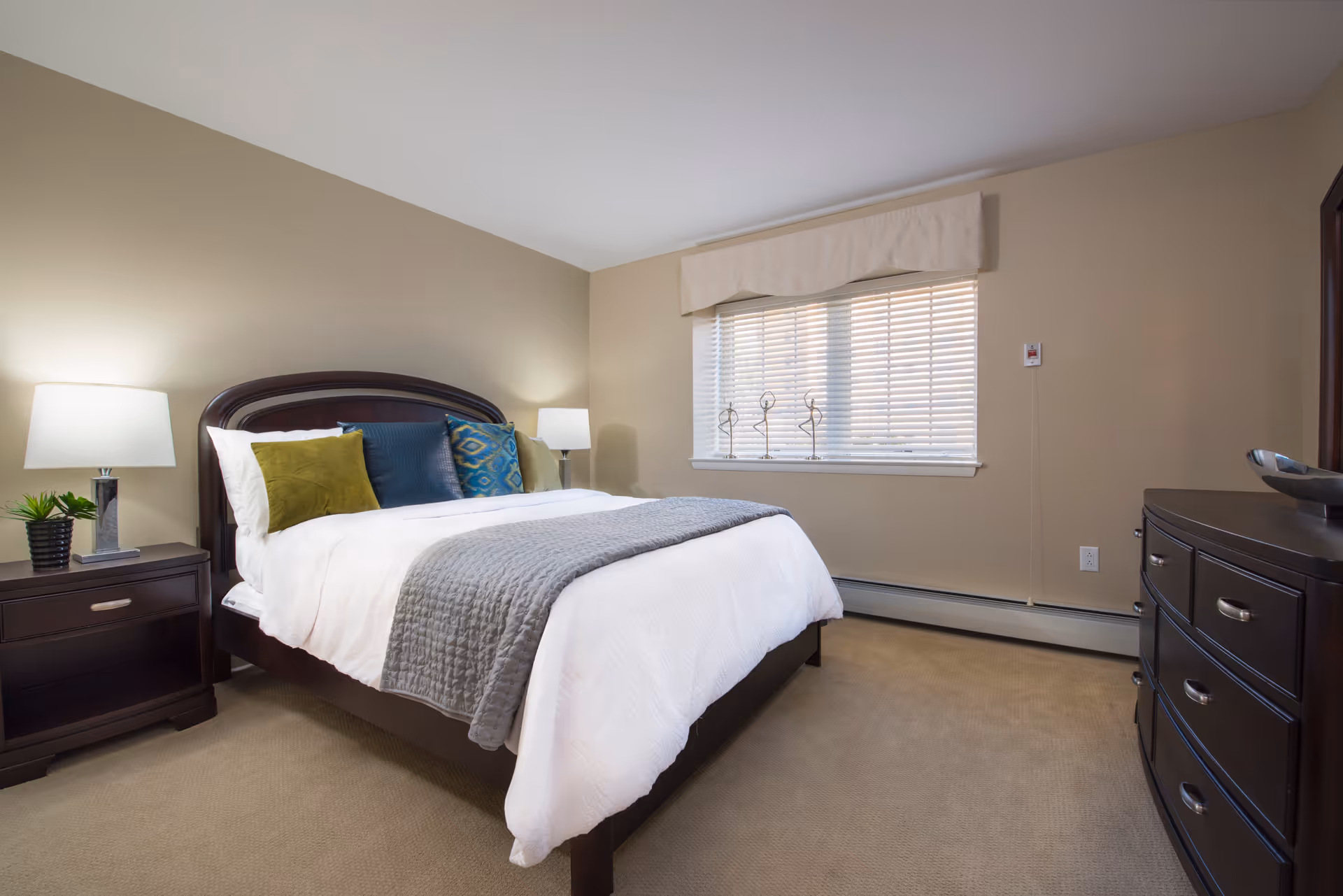 A neatly made bed with white and gray bedding and multiple decorative pillows in a bedroom. There are two bedside tables with lamps and a small plant on one side. A window with blinds and decorative items on the sill is on the wall behind the bed. A dark wooden dresser with a decorative bowl is on the right side of the room. The walls are painted beige and the floor is carpeted.
