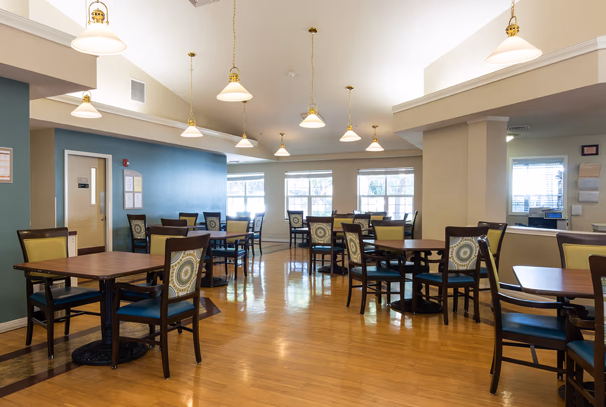 A spacious dining room in a senior living facility with multiple wooden tables and chairs arranged neatly. The chairs have patterned backs and blue seats. The room has large windows letting in natural light, wooden flooring, and several hanging pendant lights from the ceiling.
