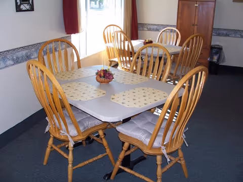 A dining area with two rectangular tables, each surrounded by four wooden chairs with cushioned seats. The tables have placemats and small floral centerpieces. The room has carpeted flooring, light-colored walls with a decorative border, and a wooden cabinet against the far wall.
