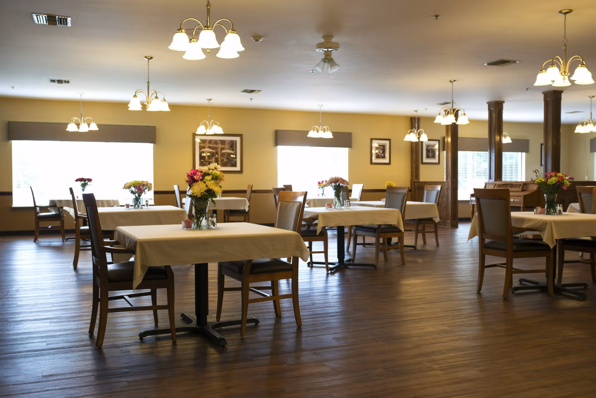 Spacious dining room with multiple tables covered in white tablecloths, wooden chairs, hanging light fixtures, and floral centerpieces.