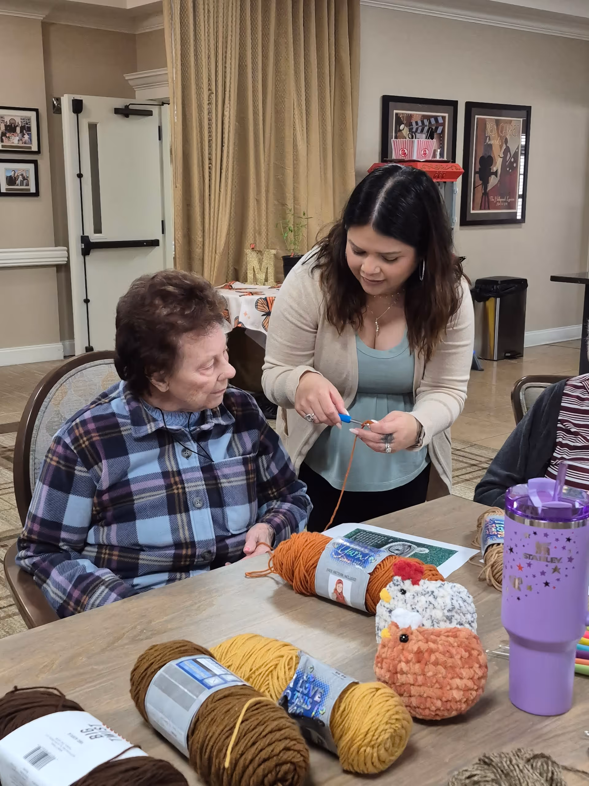 An elderly woman sitting at a table with yarn and crocheted items while a younger woman stands beside her, demonstrating or helping with a crochet project in a cozy indoor setting.