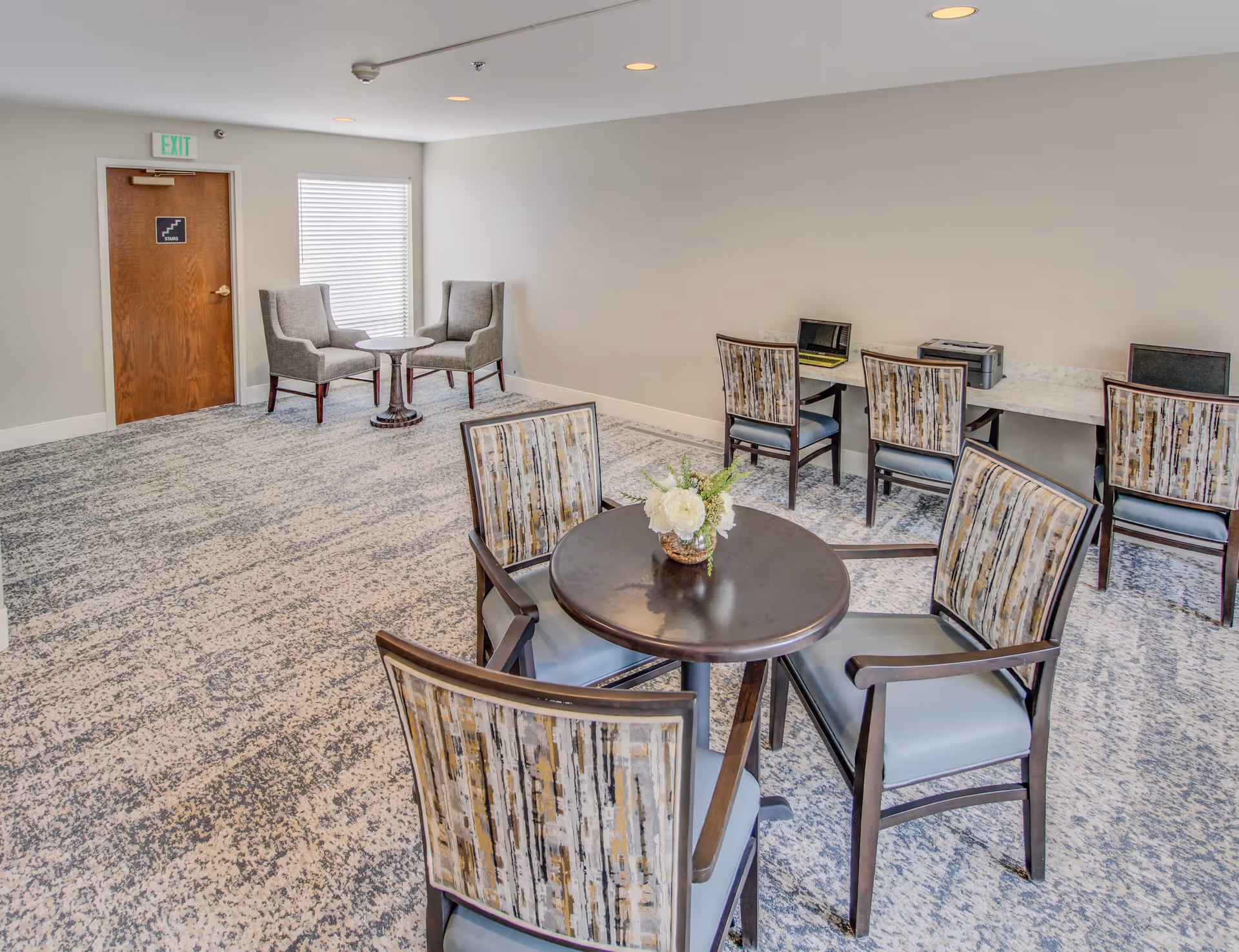 A bright room with patterned carpet featuring a round table with four chairs around it and a small flower arrangement on the table. In the background, there are two gray armchairs with a small round table between them near a window with blinds. Along one wall, there is a long desk with three chairs, a laptop, and a printer. A wooden door with an exit sign above it is visible on the left side of the room.