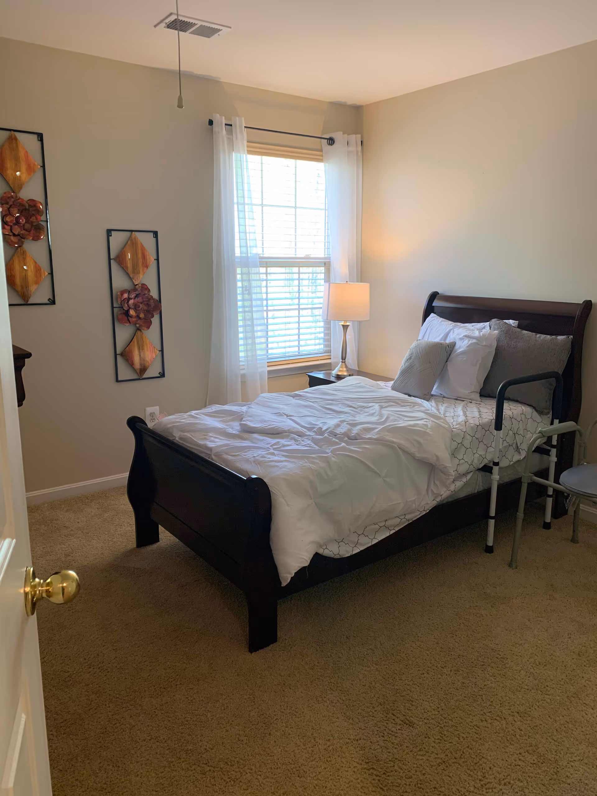 Bright bedroom with a dark wood sleigh bed made with white bedding, a bedside table and lamp, a window with sheer curtains, and a bed assist rail.