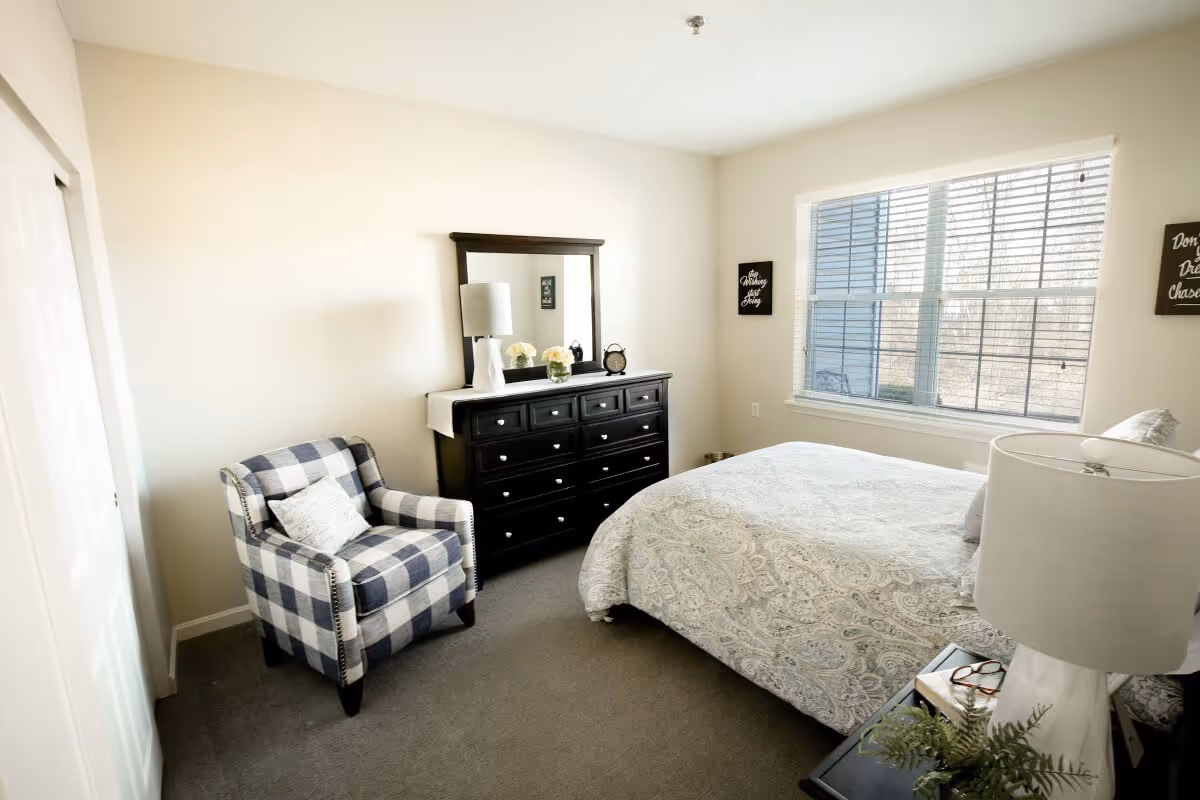 A bright bedroom with a large window letting in natural light. The room features a bed with a patterned bedspread, a black dresser with a mirror, a white table lamp, a checkered armchair with a pillow, and two small wall decorations on either side of the window.