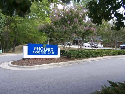 Blue Phoenix Assisted Care sign at landscaped entrance with trees and cars nearby.