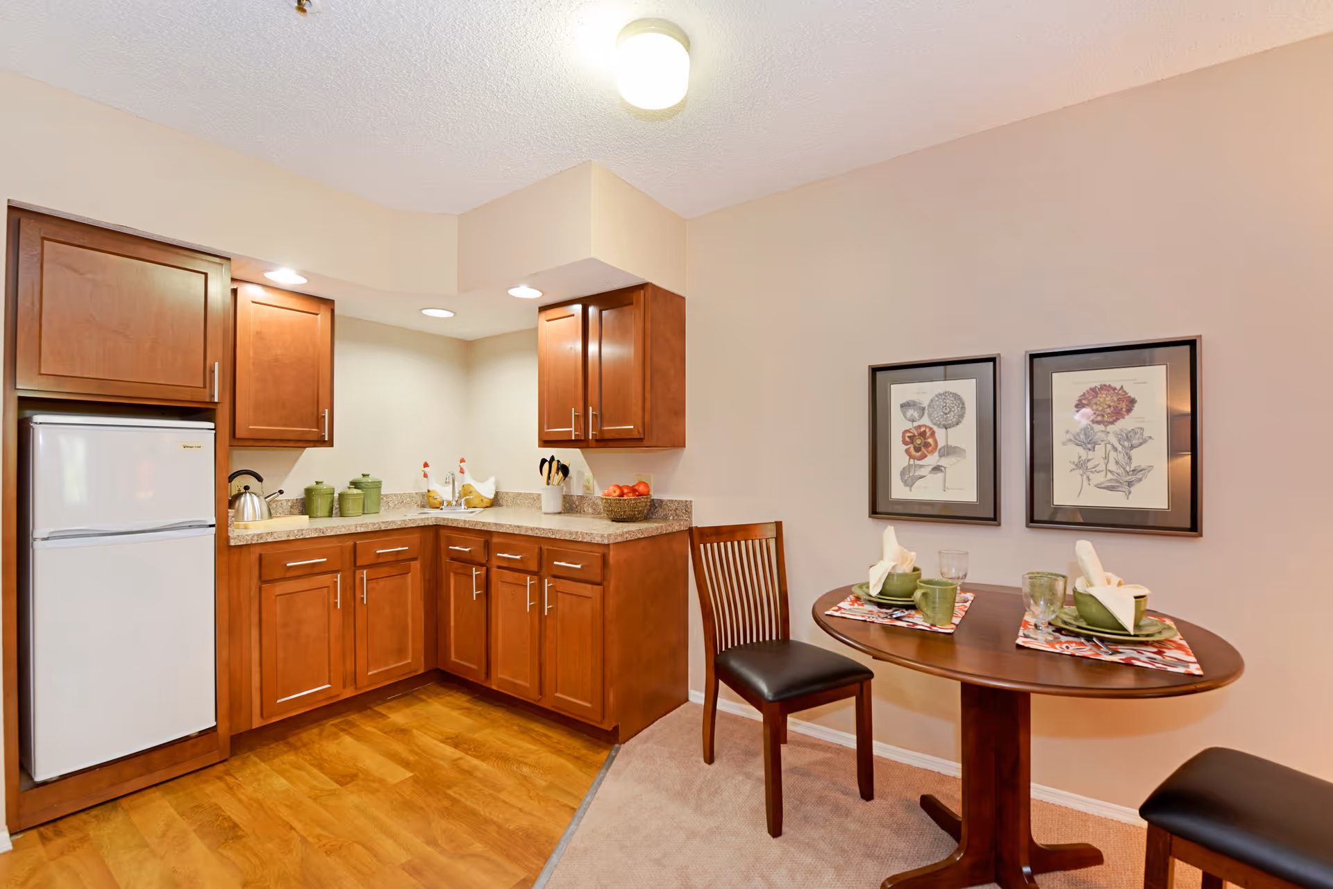 A small kitchen area with wooden cabinets, a white refrigerator, a countertop with a sink, and various kitchen items including a kettle and green canisters. Adjacent to the kitchen is a small dining area with a round wooden table set for two, including plates, cups, napkins, and placemats. Two framed floral prints hang on the wall above the dining table.