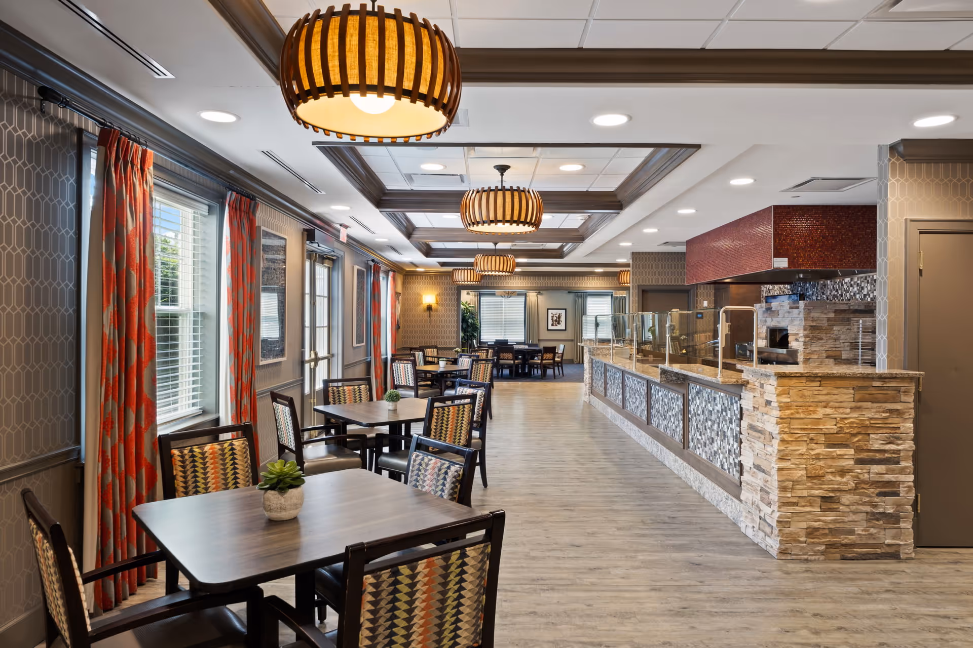 Spacious dining area with tables and chairs, pendant lights, a long serving counter with stone facade, and large windows with red curtains.