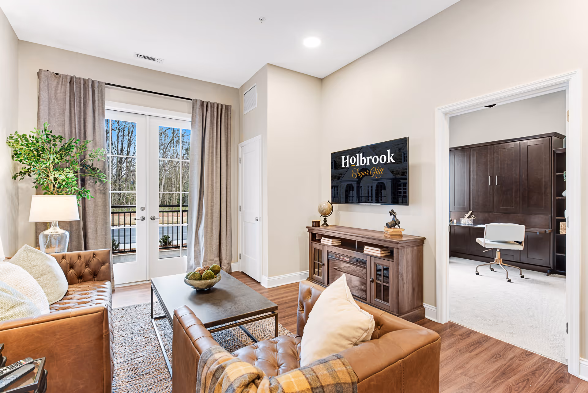 A cozy living room with two brown leather tufted sofas, a rectangular coffee table with a decorative bowl, a floor lamp with a glass base, and a large potted plant. French doors with beige curtains open to an outdoor view of trees. A wooden media console holds decorative items and a TV mounted on the wall displays the text 'Holbrook Sugar Hill'. An adjacent room with dark wood cabinetry and a white office chair is visible through an open doorway.