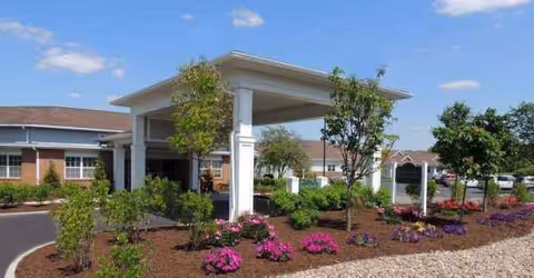 Entrance area of Laurel Lake Retirement Community featuring a covered drop-off with white columns, landscaped garden beds with green shrubs and colorful flowers, and a clear blue sky.