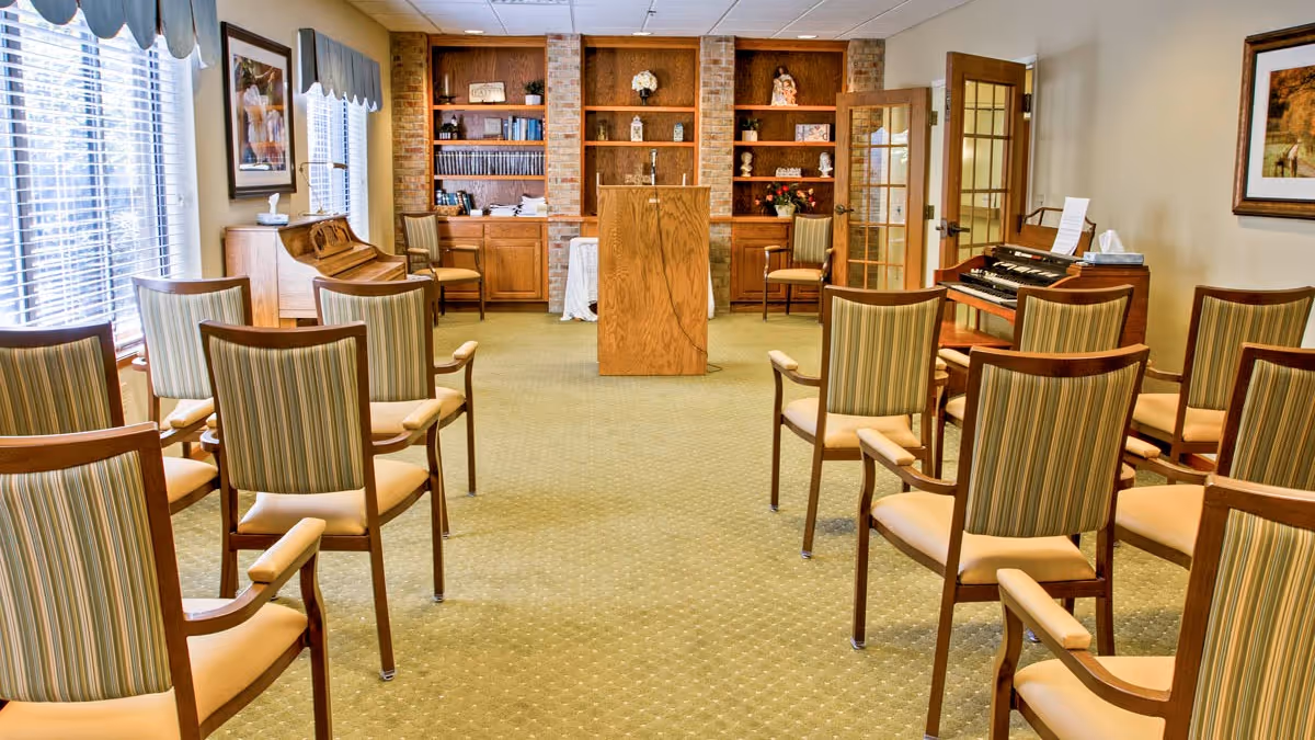 Sunlit senior living activity room with rows of striped chairs facing a wooden podium, bookshelves, a piano and an organ.