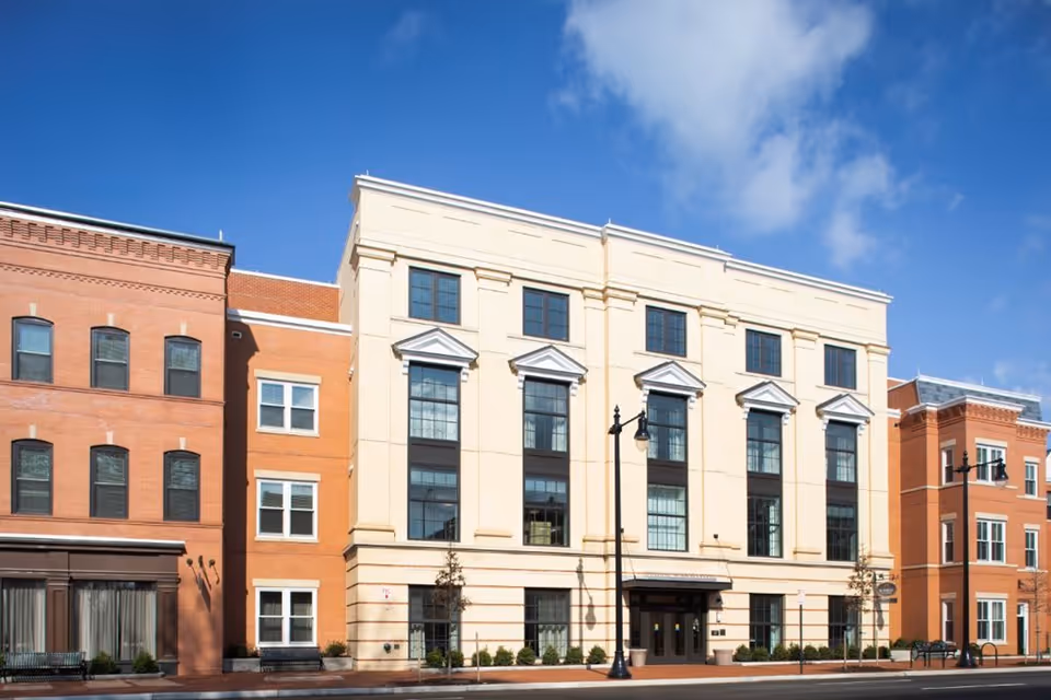Front exterior of a multi-story senior living building with brick and pale stone facades, large windows, street lamps and a blue sky.