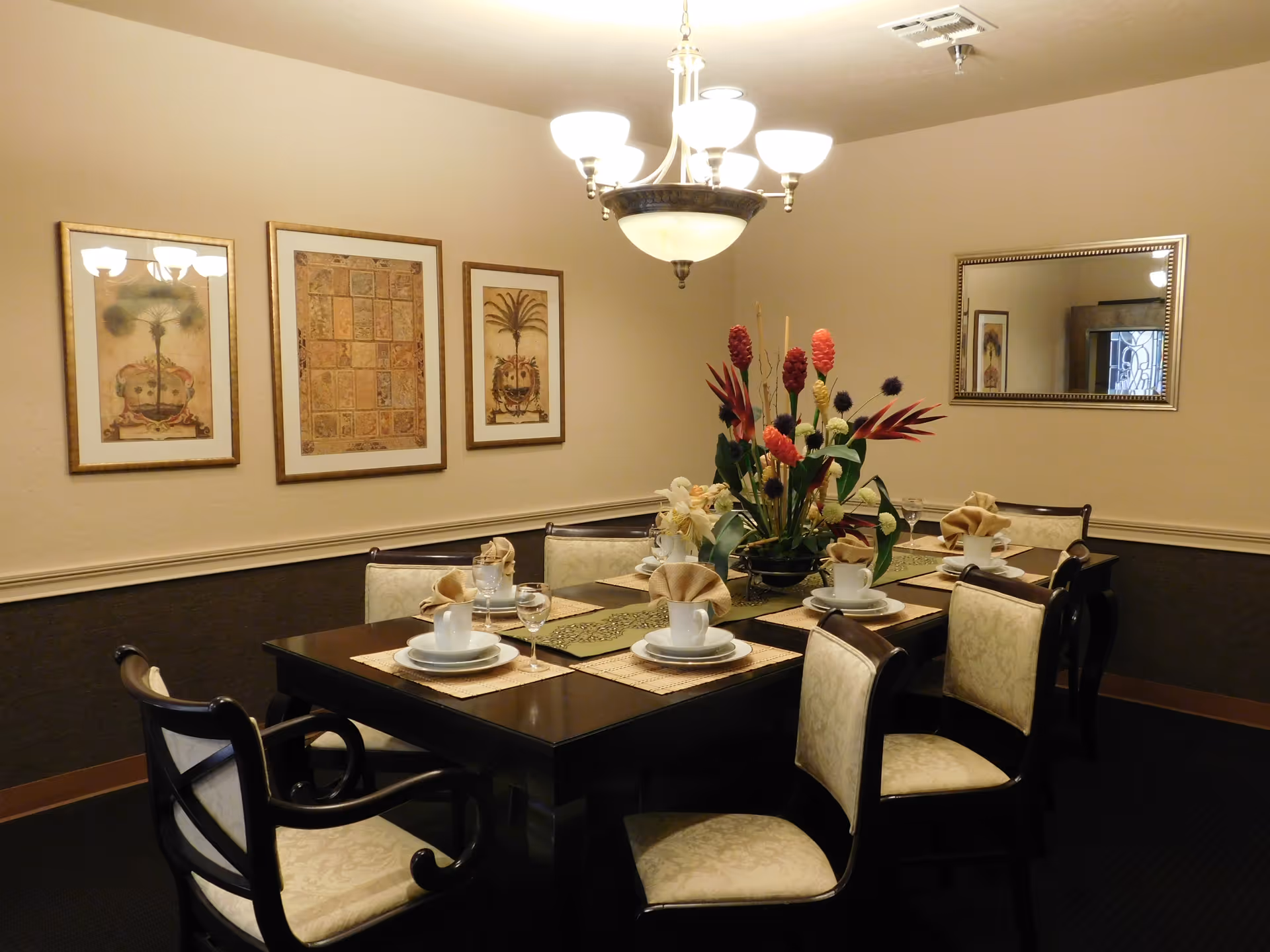 A formal dining room with a dark wooden table set for six people, each place setting including a white cup, saucer, plate, and a folded beige napkin. A floral centerpiece with red and cream flowers is in the middle of the table. The room has beige walls with three framed pictures on one wall and a large mirror on another. A chandelier with multiple lights hangs above the table.