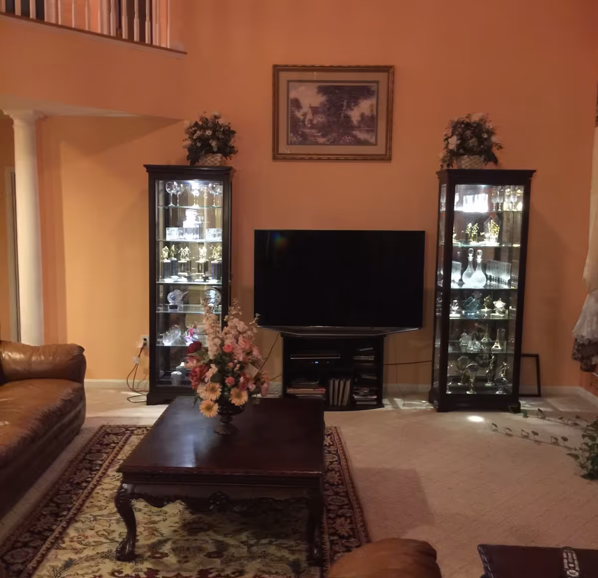 A living room with a flat-screen TV flanked by two lit glass display cabinets, a wooden coffee table with a floral arrangement, and leather seating.