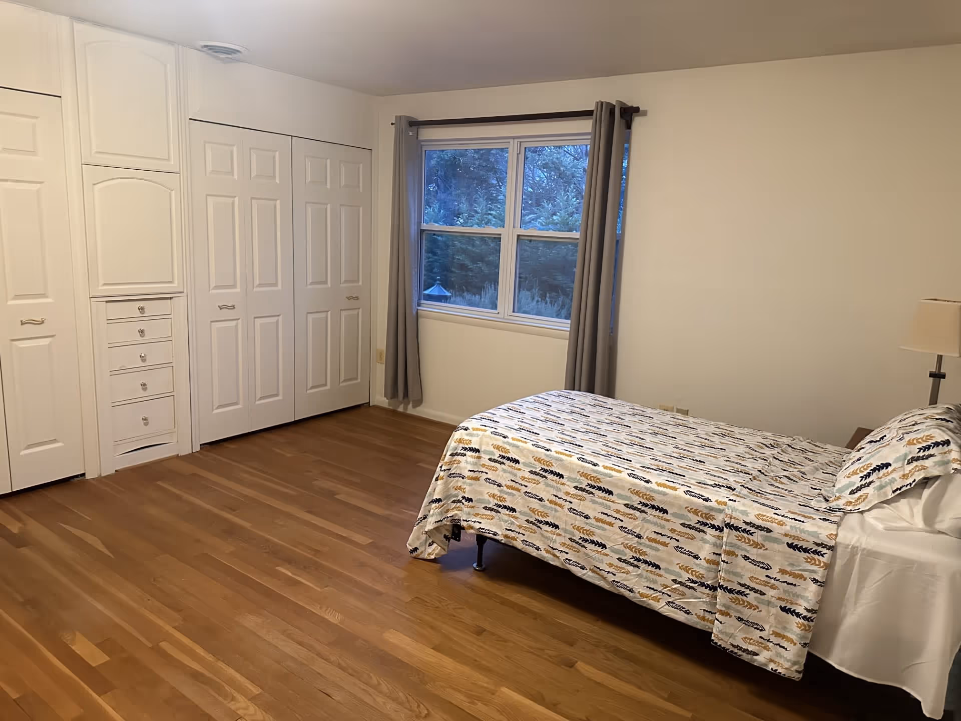 A bedroom with a wooden floor, a bed with a patterned bedspread and matching pillow, a window with gray curtains, a bedside table with a lamp, and white closets with multiple doors and drawers.