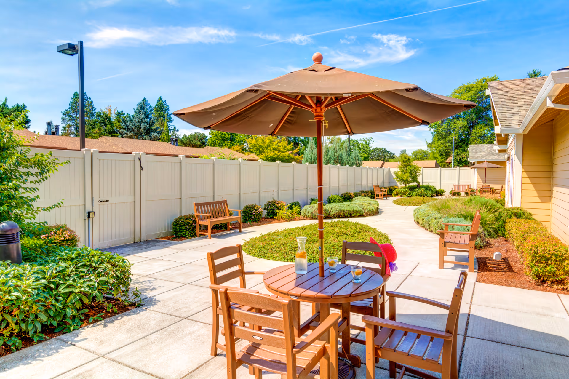 Outdoor patio area at Washington Gardens Memory Care featuring a round wooden table with four chairs and a large umbrella. The patio is surrounded by well-maintained bushes, plants, and a white fence. Additional benches and tables with umbrellas are visible along the paved walkway under a clear blue sky.