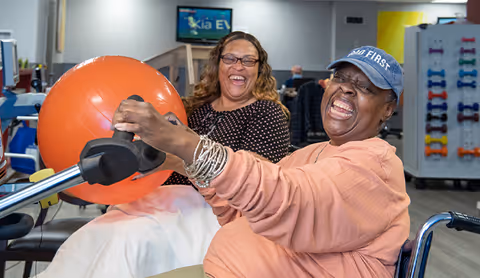 Two women smiling and laughing in a rehabilitation center. One woman in a wheelchair is holding an exercise ball attached to a piece of equipment, while the other woman sits nearby, also smiling. The background shows exercise equipment and a television screen.