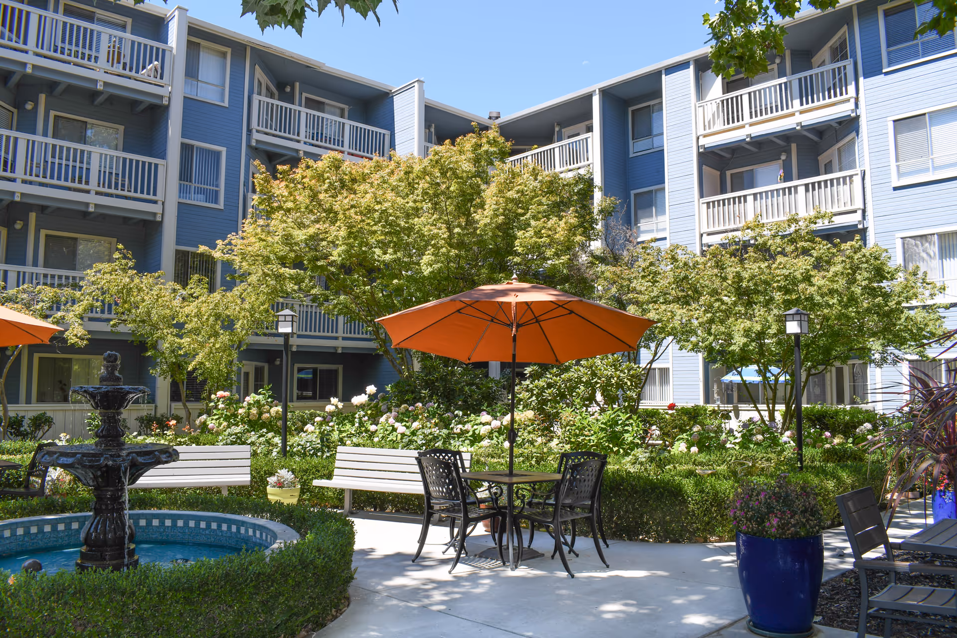 Outdoor courtyard area of a senior living facility with a blue multi-story building in the background. The courtyard features a round water fountain surrounded by greenery, white benches, a table with four black chairs, and an orange umbrella providing shade. There are trees and flowering plants around the seating area under a clear blue sky.
