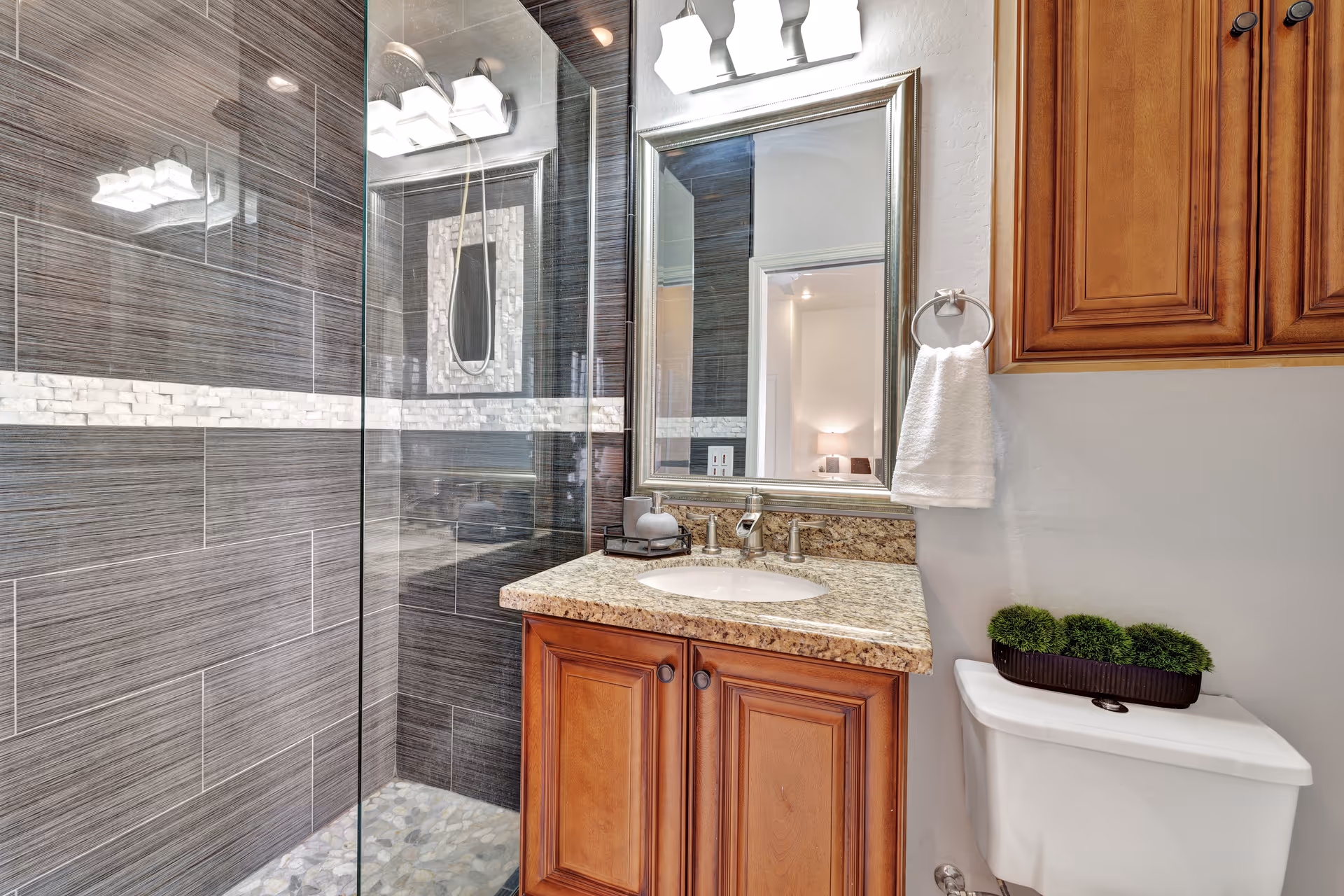 A modern bathroom featuring a glass-enclosed shower with dark gray tiles and a decorative white tile strip. There is a wooden vanity with a granite countertop, an oval sink, and a large framed mirror above it. A white towel hangs on a ring next to the mirror, and a small green plant decorates the top of the white toilet.