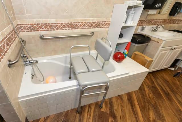 Accessible bathroom showing a bathtub with grab bars and a shower transfer chair next to a sink and shelving.