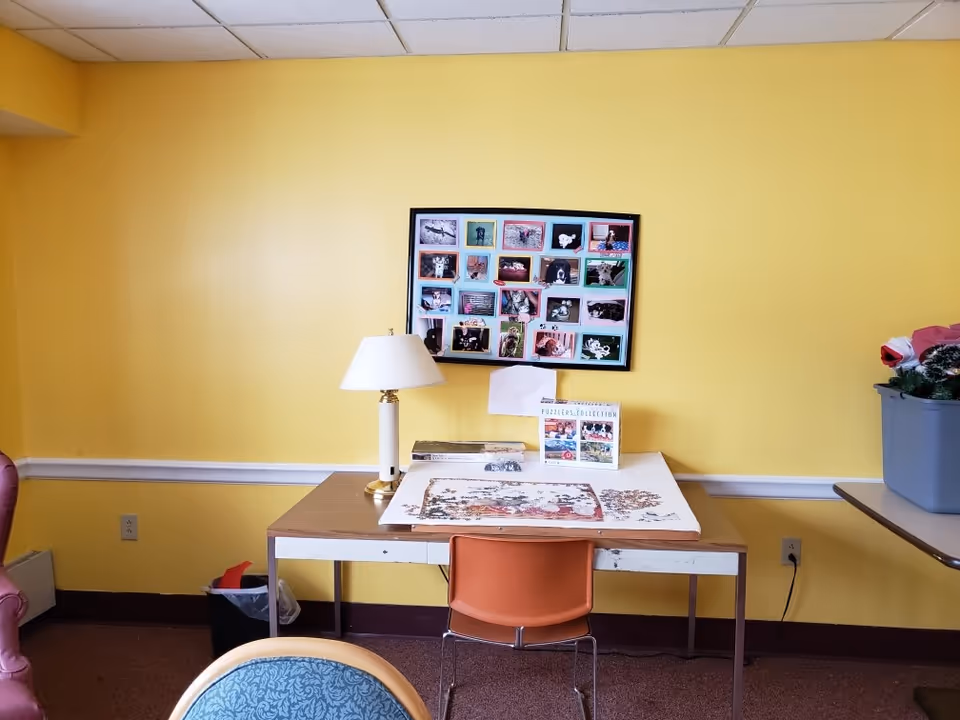 A yellow-walled interior room with a table holding a lamp and a partially completed puzzle beneath a framed collage on the wall.