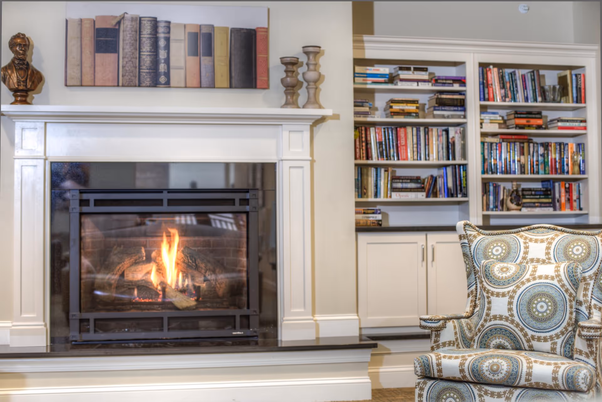 Cozy living room area with a lit fireplace surrounded by a white mantel. Above the mantel is a decorative piece featuring books and a small bust sculpture. To the right, there is a built-in bookshelf filled with various books and a patterned armchair in the foreground.