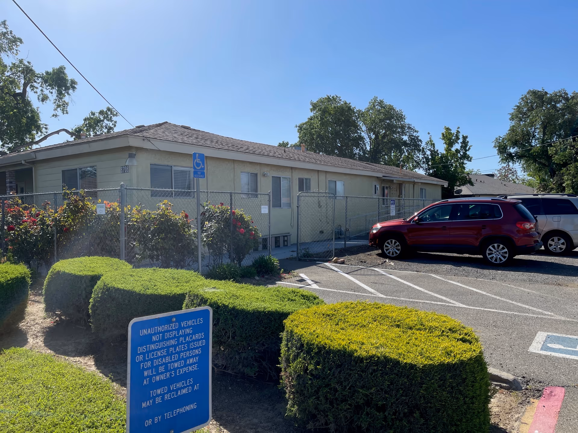 Single-story facility building with parked cars, a handicap parking sign, chain-link fence, and trimmed bushes in front.