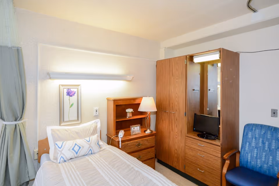 A small, neatly arranged bedroom in a senior living facility featuring a single bed with white and beige bedding, a wooden nightstand with a lamp and a small flower vase, a wooden wardrobe with mirrored doors and a small television, and a blue upholstered chair. A framed flower picture hangs on the wall above the bed, and light green curtains are partially visible on the left side.