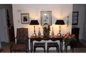 A cozy interior hallway area featuring a wooden console table with decorative items including two table lamps, a bust sculpture, and small figurines. Above the table are two framed pictures on the wall. To the left of the table is an upholstered armchair with a patterned fabric. To the right is a floral arrangement on a small stand. The space is warmly lit and has a traditional decor style.
