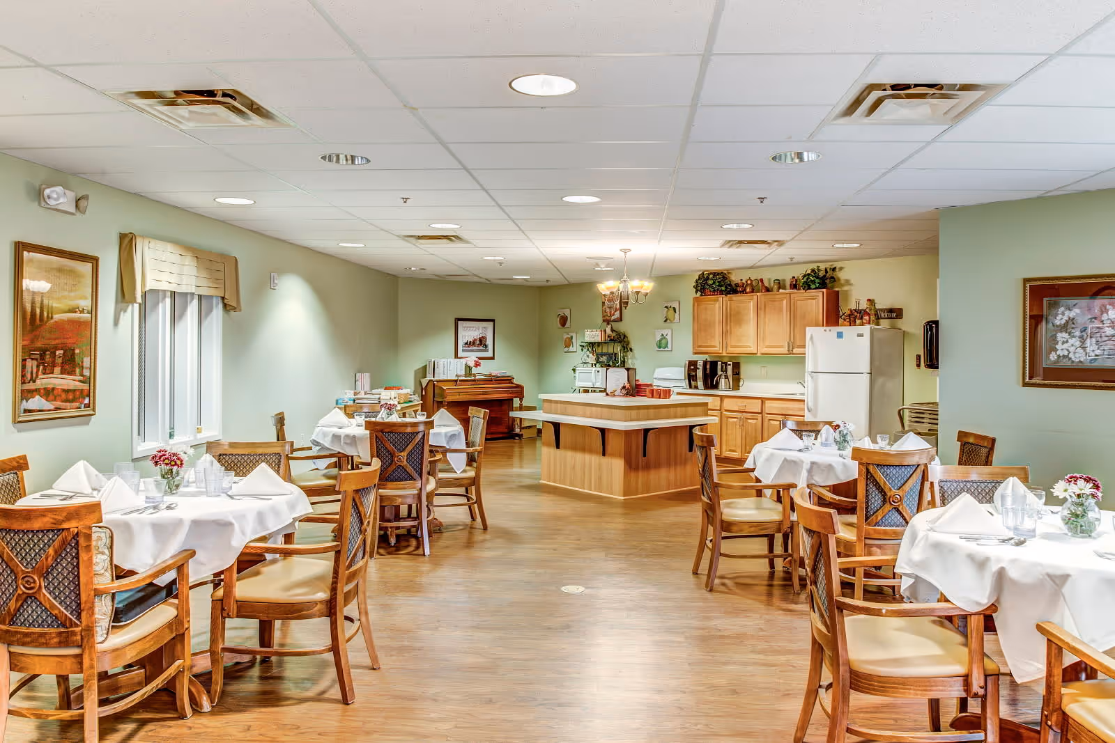 A dining room in a senior living facility with several round tables covered with white tablecloths, each set with glasses, napkins, and small flower arrangements. Wooden chairs with cushioned seats surround the tables. In the background, there is a kitchen area with wooden cabinets, a refrigerator, a microwave, and a coffee maker. The room has light green walls, framed artwork, and a wooden floor.