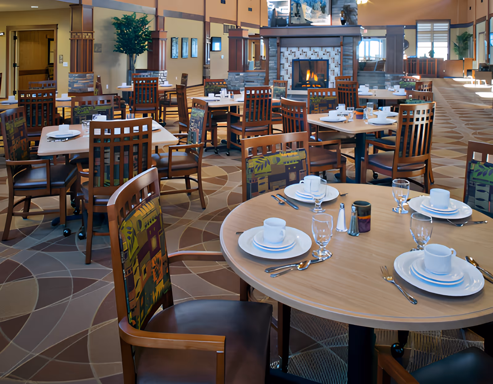 A spacious dining room with multiple round and square tables set with white plates, cups, silverware, and glasses. The chairs have wooden frames with patterned fabric backs. A fireplace with a lit fire is visible in the background, along with large windows letting in natural light and decorative plants.