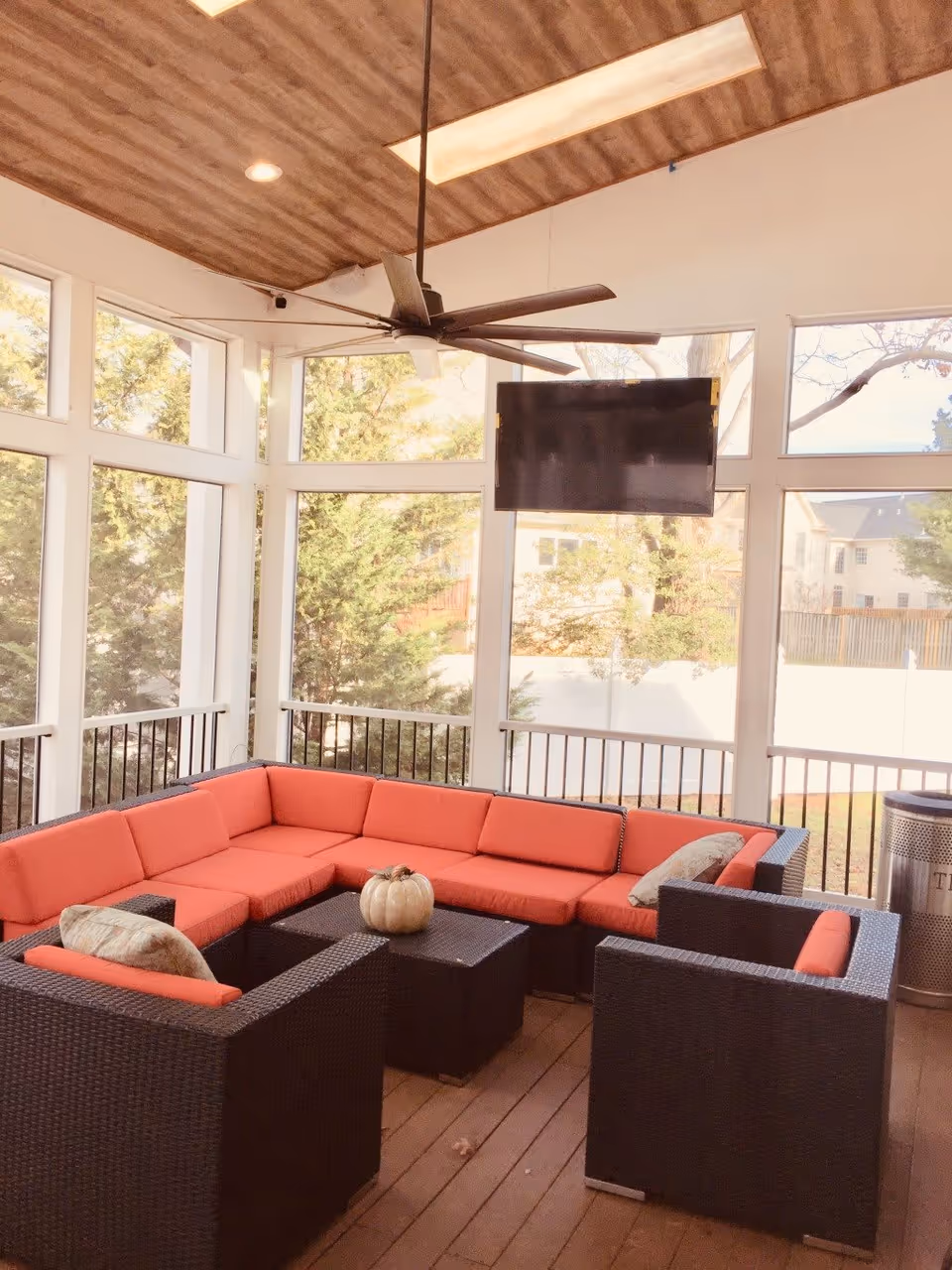 Enclosed patio lounge with orange-cushioned wicker sectional, coffee tables, a ceiling fan, and a wall-mounted TV in front of large windows.