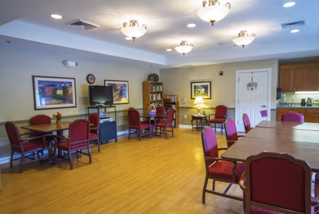 Spacious assisted-living common room with several tables and red upholstered chairs, a TV and bookshelf, and a kitchenette area.