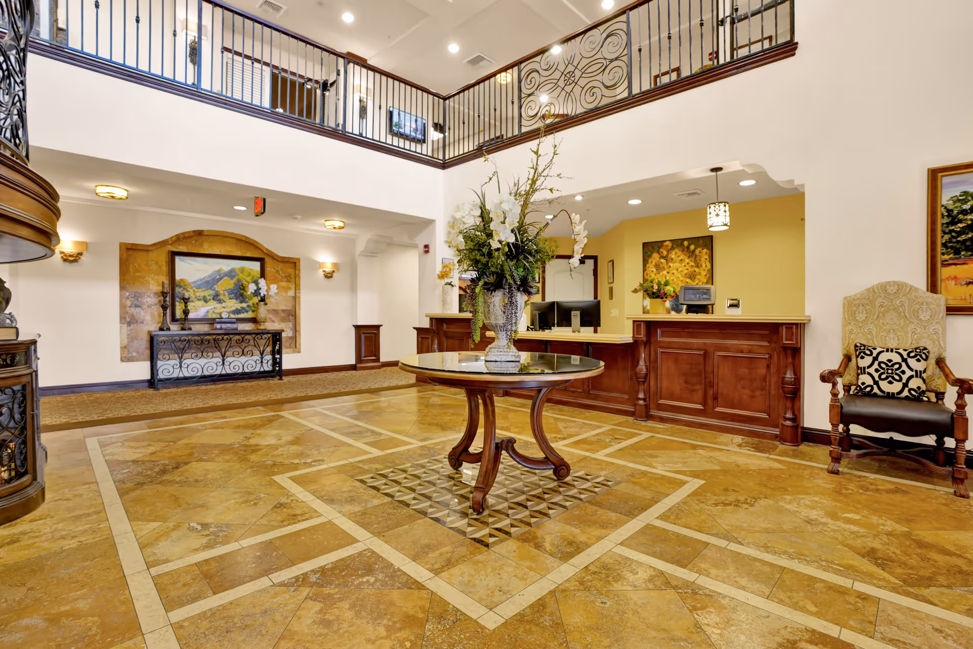 Spacious and elegant lobby area with a round wooden table topped with a large floral arrangement in the center. The floor features a decorative tile pattern. To the right is a wooden reception desk with a computer and a chair with a patterned cushion. The walls are adorned with framed artwork, and there is a second-floor balcony with wrought iron railings overlooking the lobby.
