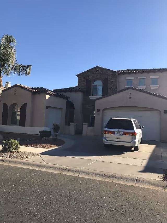 Exterior view of a two-story residential building with a beige stucco finish and a tiled roof. There is a driveway with a white minivan parked in front of a double garage door. The building has arched windows with dark shutters and a small landscaped area with desert plants and a palm tree. The sky is clear and blue.