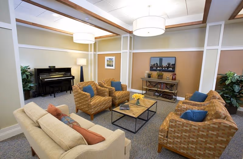 A cozy living room area in an assisted living facility featuring a beige sofa with red and patterned cushions, four patterned armchairs with blue cushions arranged around two square wooden coffee tables with a vase of yellow flowers, a black upright piano in the corner, a floor lamp, a TV on a console table, and decorative plants. The room has soft lighting from ceiling fixtures and a neutral color palette with beige and light brown walls.
