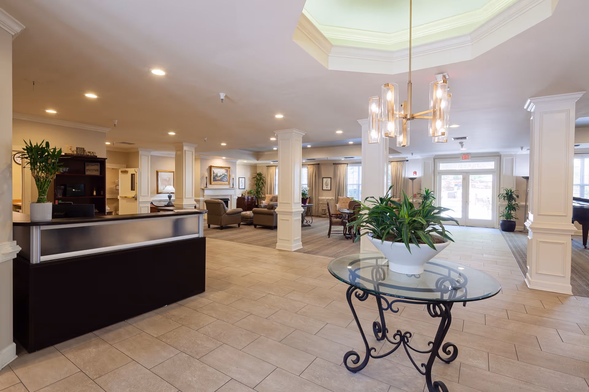 A spacious and well-lit senior living facility lobby with a reception desk on the left, a glass table with a potted plant in the center, and comfortable seating areas with armchairs and sofas in the background. The room features tiled flooring, white pillars, and large windows allowing natural light to enter.