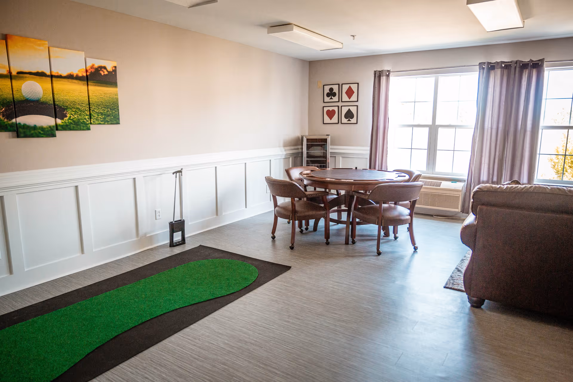 Bright common room with a round table and chairs, a small putting green mat, sofa, large window with curtains, and wall art.