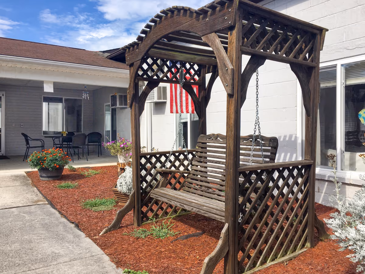 A wooden outdoor swing with a lattice design and a canopy, placed on a mulched garden bed with some small plants. Behind the swing is a white building wall with windows and an American flag hanging. To the left, there is a covered patio area with a table and chairs.