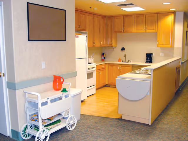Interior view of a kitchen area in a senior living facility with wooden cabinets, a white refrigerator, stove, and coffee maker on the counter. A white utility cart with a red pitcher and green cup is positioned near the entrance. The floor transitions from carpet in the hallway to wood in the kitchen.