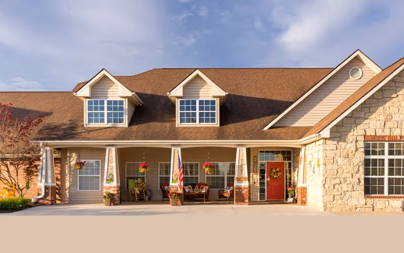 Front exterior view of a single-story residential style building with a brown shingled roof, beige siding, and stone accents. The entrance features a covered porch with white columns, hanging flower baskets, an American flag, and a red door decorated with a wreath. There are large windows on the front facade and some outdoor seating on the porch.